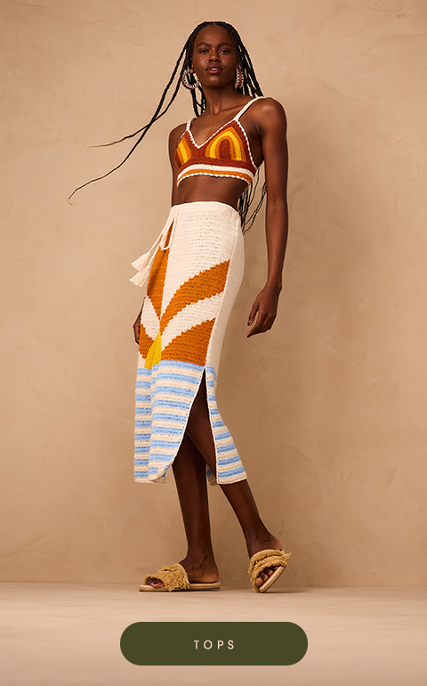 Woman wearing a sleeveless brown top with a white diamond pattern and side ties, paired with high-waisted cream pants. She is smiling and standing against a beige textured background.