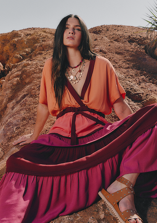  A brunette model is sitting on a rock, wearing a long-sleeve dress with a V-neck in wine, pink, and orange.