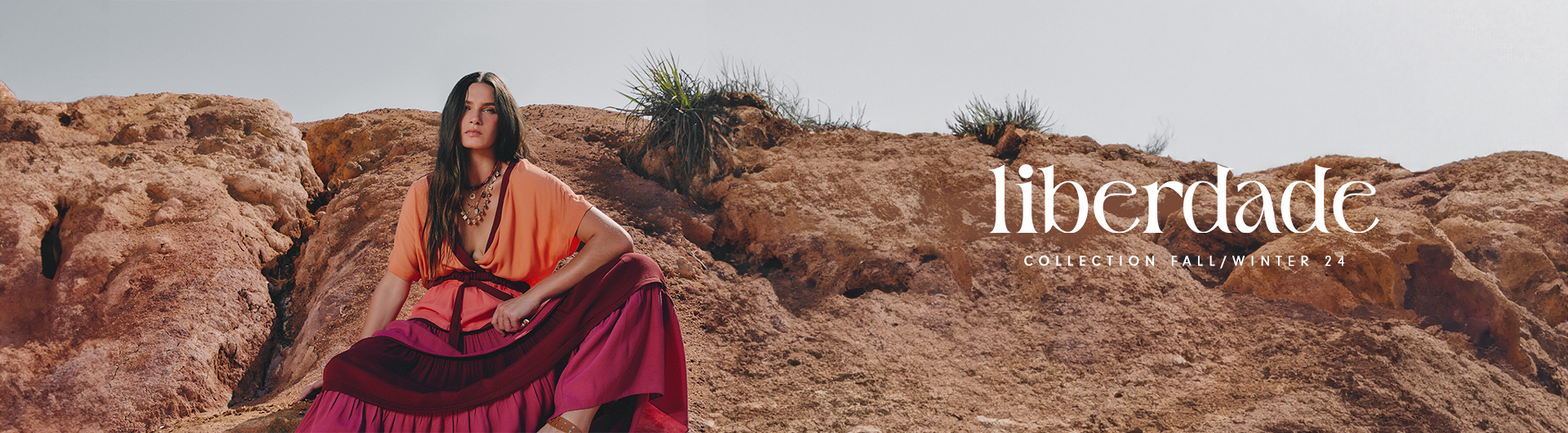 A brunette model with long brown hair is wearing a long-sleeved dress with a V-neck in shades of orange, pink, and wine. Complementing the look, the model wears caramel-colored sandals with silver details and accessorizes with gold necklaces and beaded heart charms. The model is seated on a sandy stone ground with a blue sky backdrop, posing for the winter collection campaign banner of the Rio de Janeiro brand Dress To.