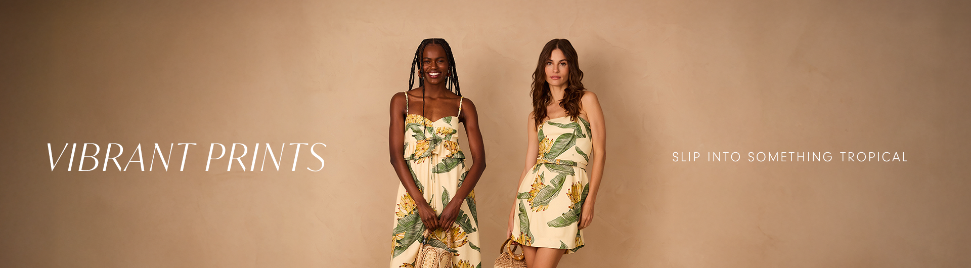 Two women wearing tropical-print dresses with green leaves and yellow flowers, standing against a warm beige background. One wears a long flowy dress, the other a short dress with a woven handbag. Text reads: 'Vibrant Prints – Slip into something tropical.'