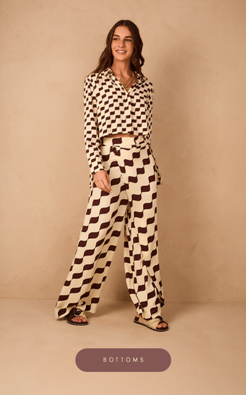 Woman wearing a matching set with a wavy checkered pattern in cream and dark brown. The outfit includes a long-sleeved button-up shirt and high-waisted wide-leg pants. She is standing against a beige textured wall, smiling, and wearing flat sandals.