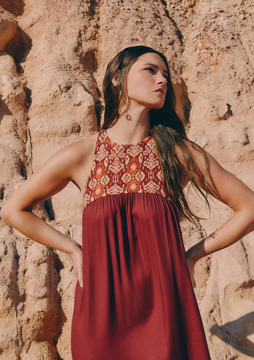 A blonde model is wearing a red dress with a printed detail on the bust and wine-colored sandals.