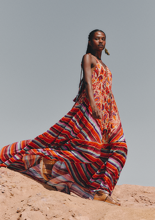 A Black model is wearing a long, warm-toned printed dress paired with beige sandals. The model is set against a backdrop of blue sky and sand dunes.