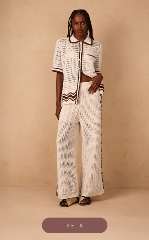 Model wearing a white crochet two-piece set from Dress To – Rio de Janeiro Collection, consisting of a short-sleeve collared button-up shirt and wide-leg pants with brown zigzag details on the sides. She stands confidently against a beige textured background. At the bottom center, there is a mauve button with white text that reads 'SETS'.