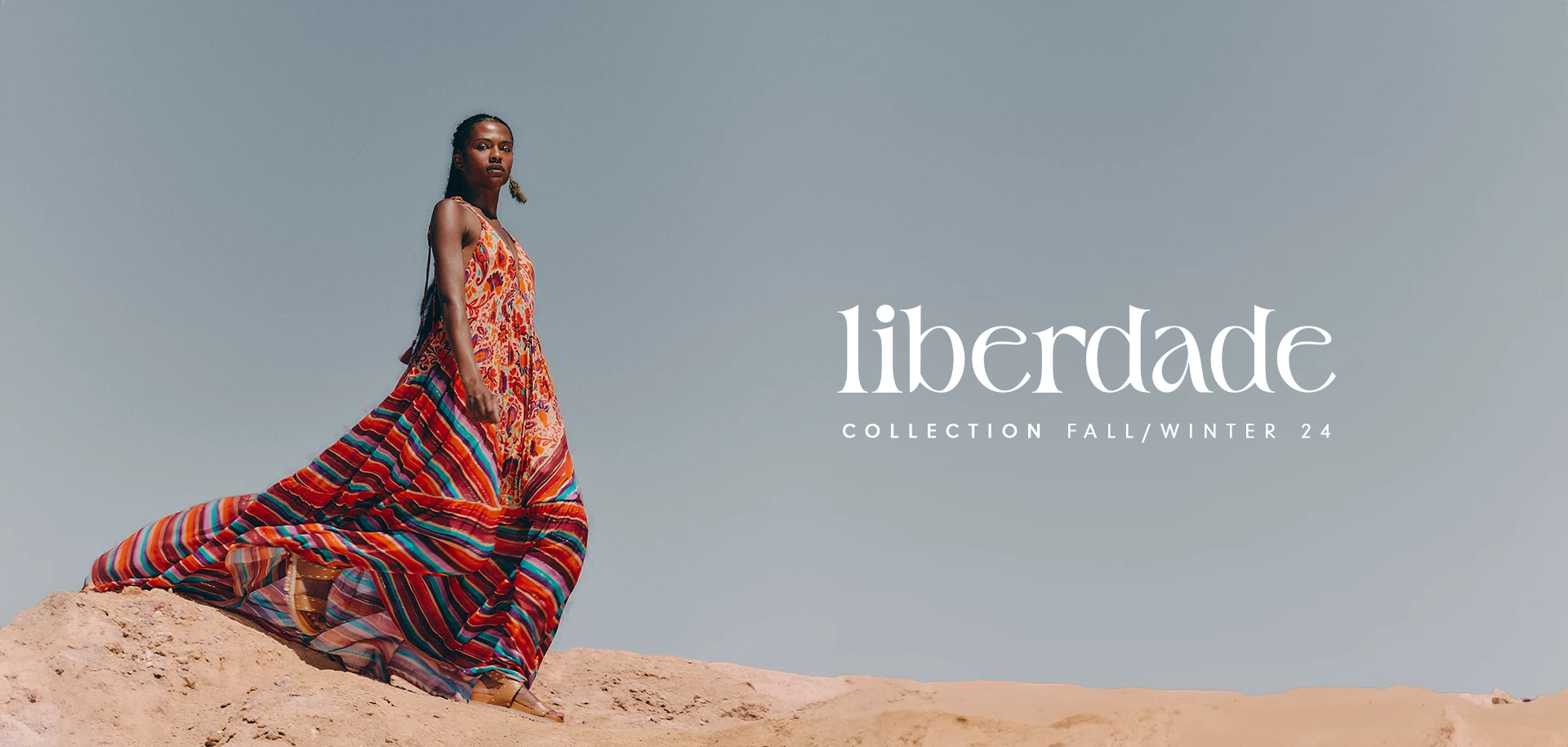 Black model wearing a long dress with the Mantra Mix print in orange, red, blue, lilac, and beige. Model featured in a campaign banner for the Liberty Collection, with a background of stone and sand dunes.