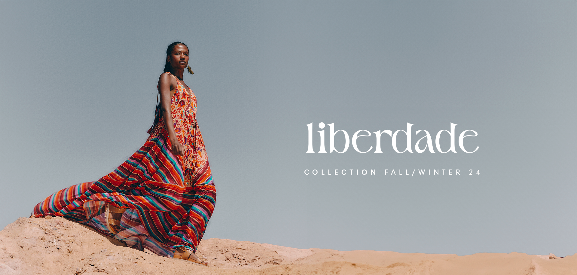 Black model wearing a long dress with the Mantra Mix print in orange, red, blue, lilac, and beige. Model featured in a campaign banner for the Liberty Collection, with a background of stone and sand dunes.