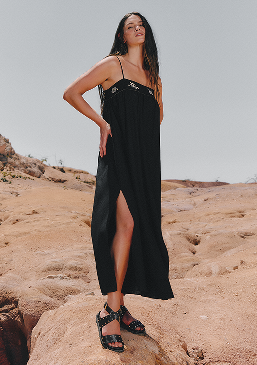 A model is wearing a black dress with embroidered details on the bust and black sandals