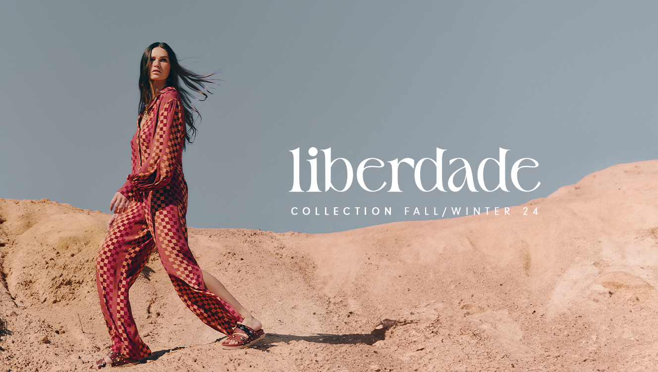 A brunette model is wearing a wine and beige square-patterned set, paired with wine-colored flat sandals from Dress To’s winter collection. The model is set against a backdrop of sand dunes with a blue sky.