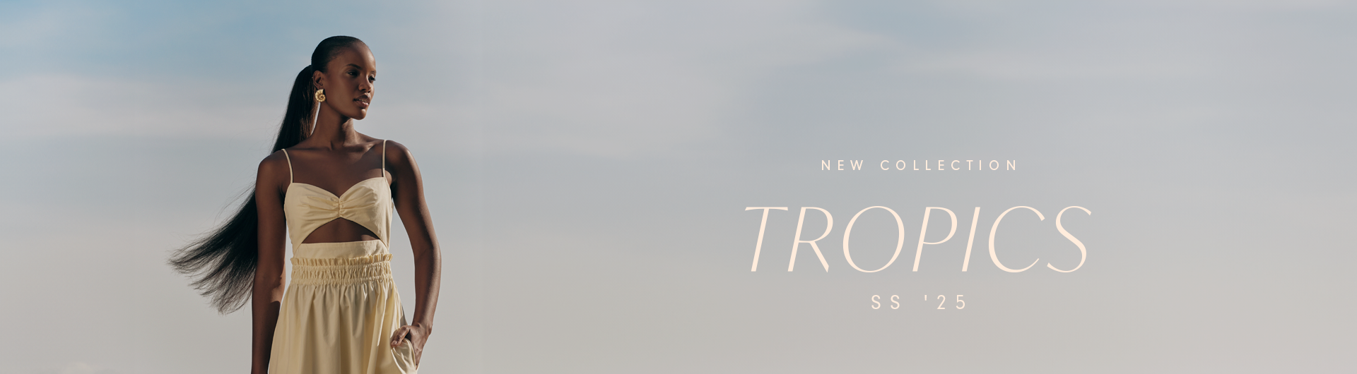 Banner for the 'TROPICS' SS '25 collection. On the left, a Black model with long, sleek hair tied in a ponytail wears a beige set featuring a thin-strapped top with a front cutout and a flowy high-waisted skirt. The background showcases a soft blue sky with subtle gradients. On the right, the text 'NEW COLLECTION TROPICS SS '25' is displayed in an elegant, minimalist font.