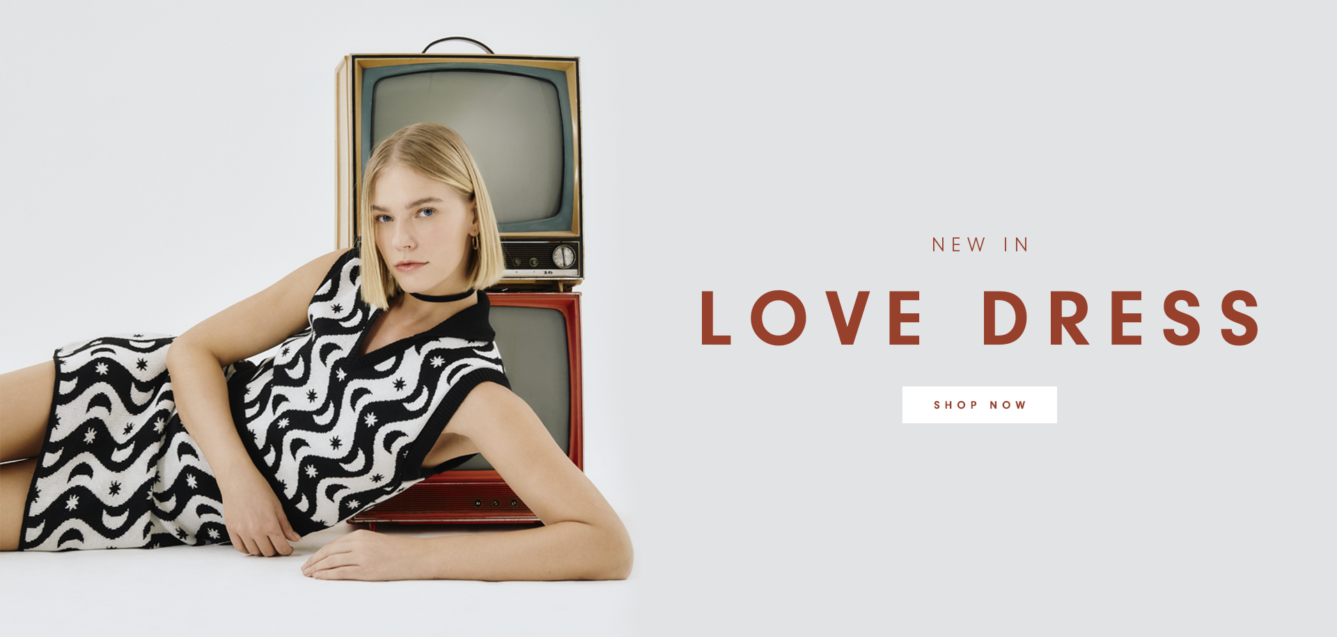 Banner with a light gray background featuring a woman with blonde hair, wearing a black and white patterned dress with wavy designs and small star motifs. She is reclining beside two vintage televisions stacked on top of each other. On the right side, the text 'NEW IN LOVE DRESS' is displayed in bold red letters.