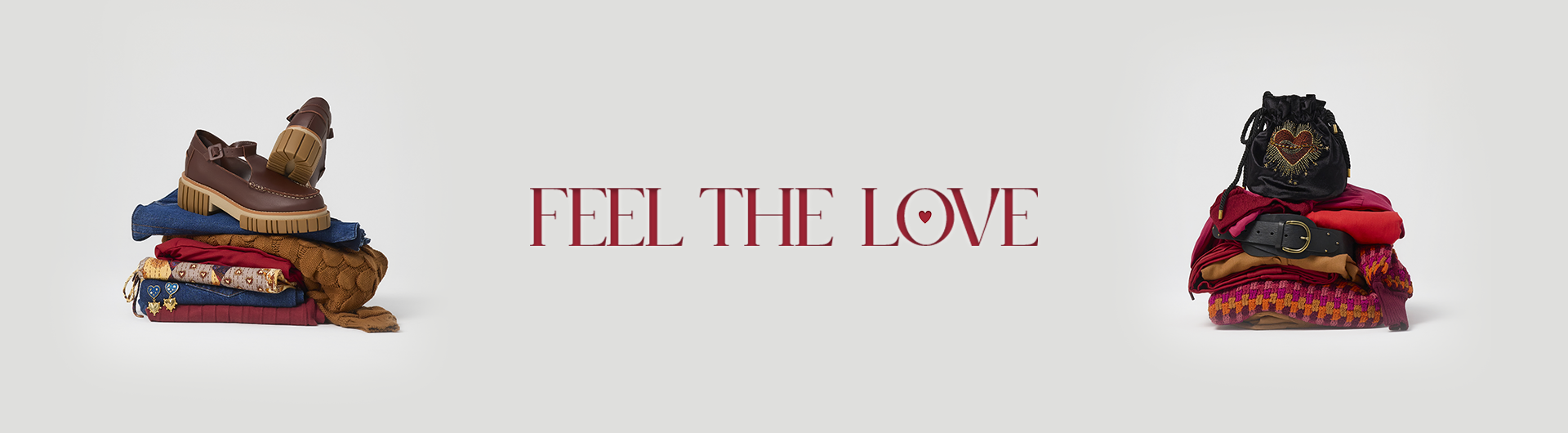 banner with a light gray background featuring the phrase "FEEL THE LOVE" in bold, red serif font, with a small heart inside the letter "O." On the left side, there is a neatly stacked pile of clothing in warm tones, including denim, knitwear, and embroidered fabrics, topped with a pair of brown chunky-sole loafers. On the right side, another stack of clothing in red, orange, and brown hues is accessorized with a black belt and a black drawstring bag adorned with a beaded heart design. The aesthetic conveys warmth, love, and a cozy fashion theme.