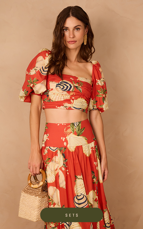 Woman wearing a cream crochet set with brown details, including a short-sleeve button-up shirt and wide-leg pants. She poses confidently against a beige textured background, wearing flat sandals.