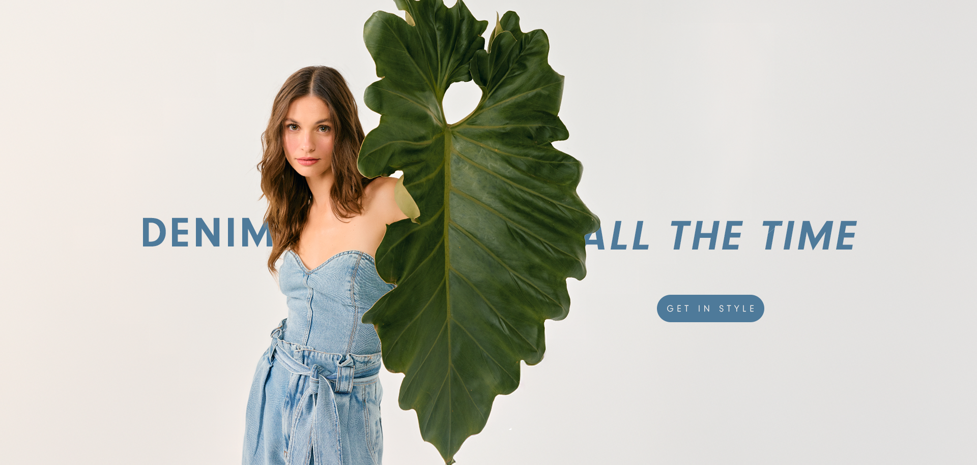Woman with loose brown hair wearing a strapless denim jumpsuit with a waist tie, partially covered by a large green leaf. Text reads: “DENIM ALL THE TIME.” Button: “GET IN STYLE.”