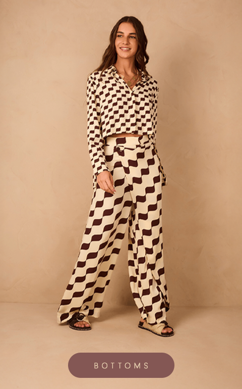 Model dressed in a wide-leg pants and cropped shirt set from Dress To – Rio de Janeiro Collection, with a geometric wavy pattern in black and cream. The outfit is paired with black sandals. She stands smiling against a light beige textured wall. At the bottom center, there is a mauve button with white text that reads 'BOTTOMS'.