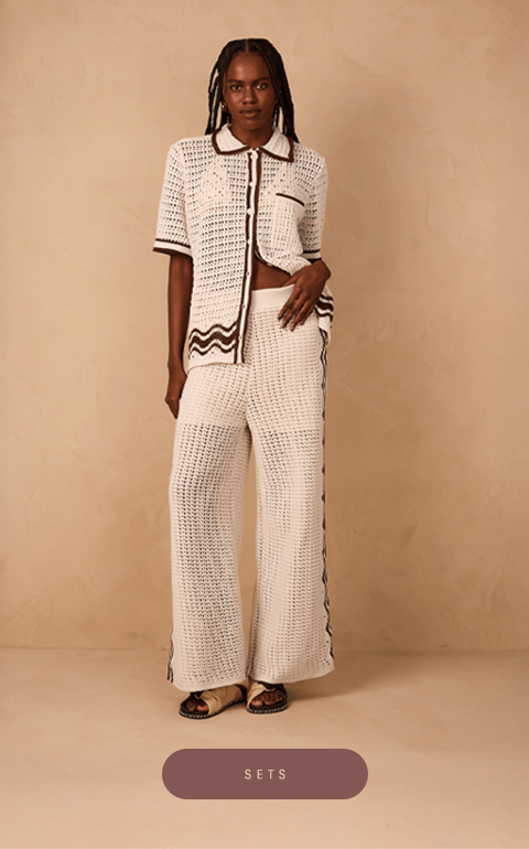 Woman wearing a cream crochet set with brown details, including a short-sleeve button-up shirt and wide-leg pants. She poses confidently against a beige textured background, wearing flat sandals.