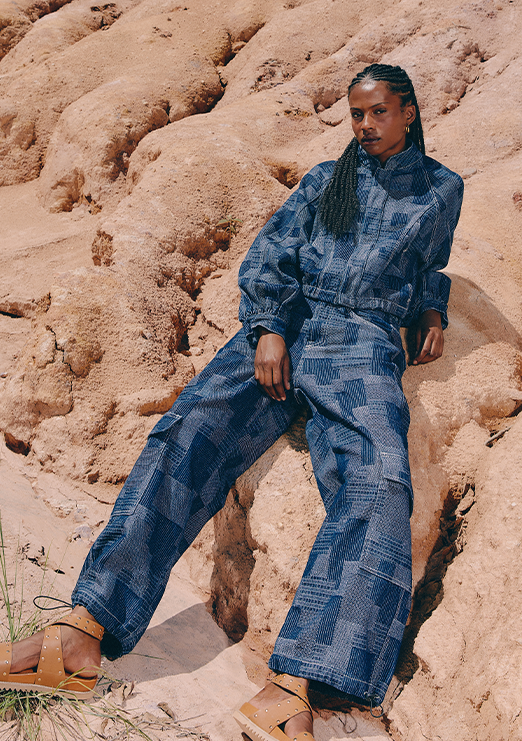  A Black model is wearing a denim set with darker checkered details, leaning against rocks in shades of brown.